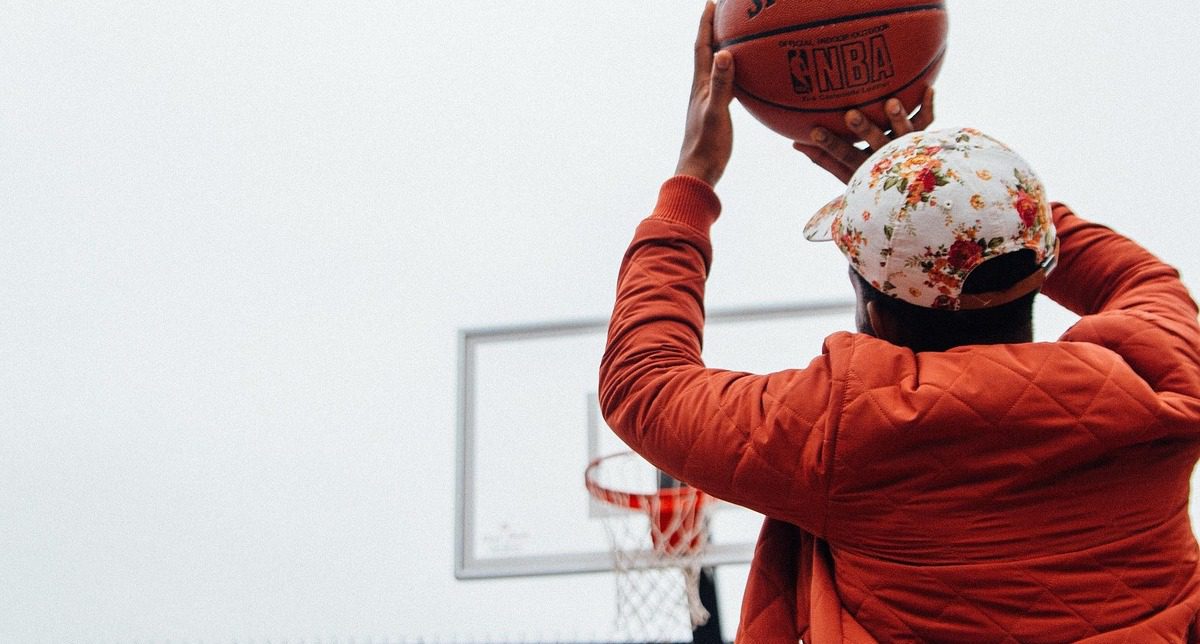 Man in an orange jacket and a floral baseball cap shooting a basketball